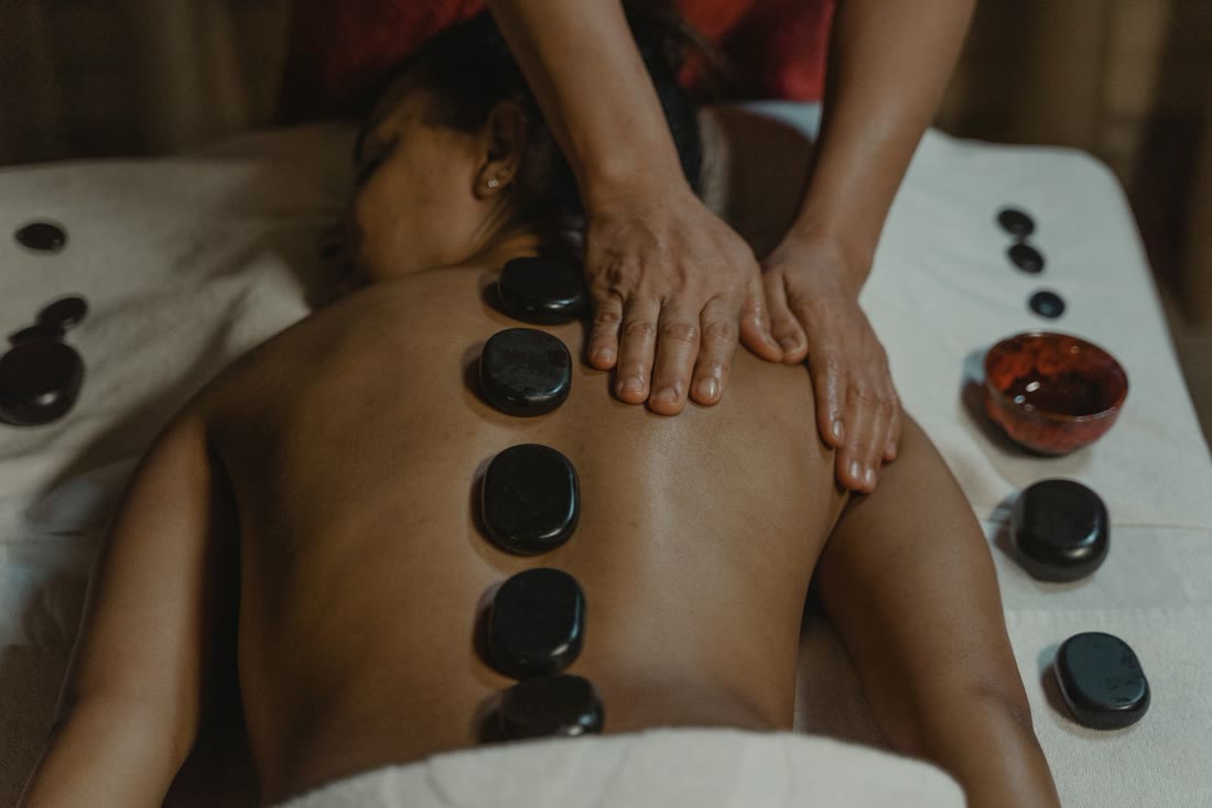 Massage aux pierres chaudes : les mains du praticien travaillent le dos, pierres de basalte alignées le long de la colonne et bol d'huile à côté
